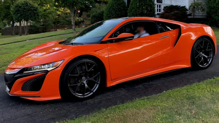 Bright orange luxury sports car with large black wheels parked on a suburban street with greenery in the background.