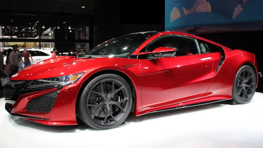 Red exotic sports car on display at an auto show, with large grey wheels and glossy red finish, crowd in the background.