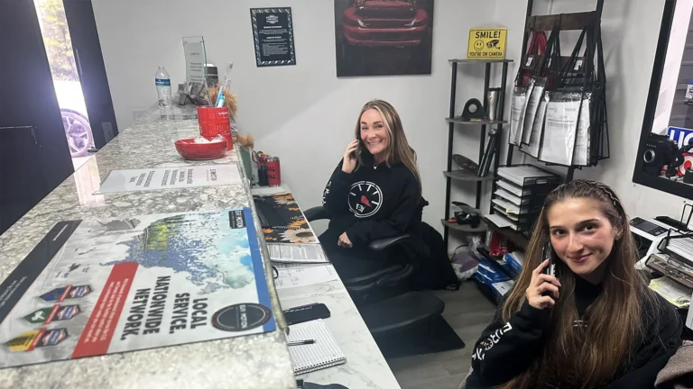 Two smiling women behind a marble reception counter, both on phones in a small office space with brochures and supplies nearby.