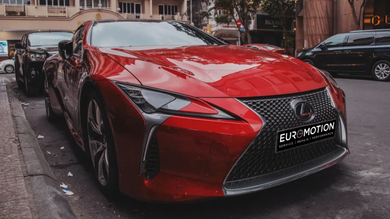 Red Lexus sports car parked on a city street with storefronts in the background and a black SUV nearby, Euro Motion plate visible on the front.